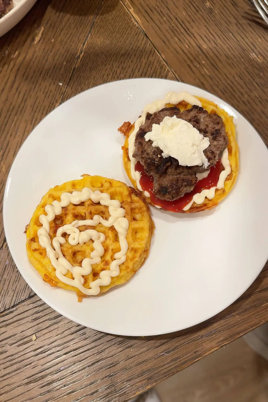 A white plate with two carnivore bread chaffles, one topped with a cooked beef patty and a swirl of mayo and ketchup, while the other is waiting to be stacked to complete the burger.